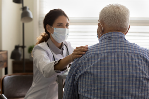 It will be all right. Empathic young female doctor in medical mask on face hold hand on shoulder of elderly man patient encourage to fight with disease. Supportive woman gp comforting old male client
